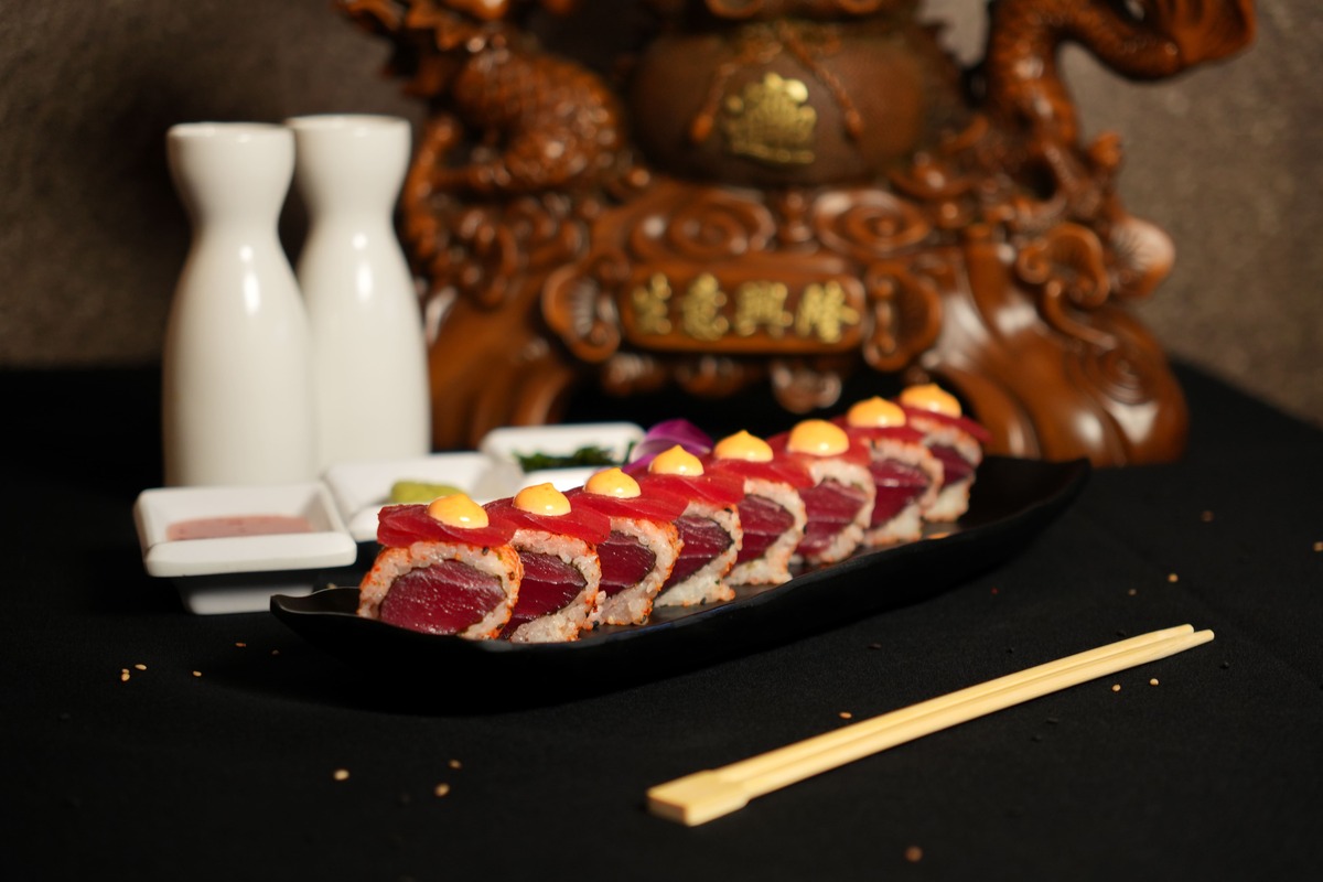 A row of sushi rolls topped with orange sauce is arranged on a black plate beside wooden chopsticks. In the background, two white sake bottles and a decorative wooden dragon sculpture evoke the elegance of the Kingdom Sushi a la carte experience.