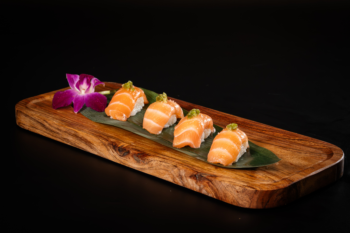 Four pieces of salmon nigiri sushi with green garnish are arranged on a green leaf atop a wooden serving tray, with a purple orchid flower for decoration—an elegant highlight from the Kingdom Sushi menu against a black background.
