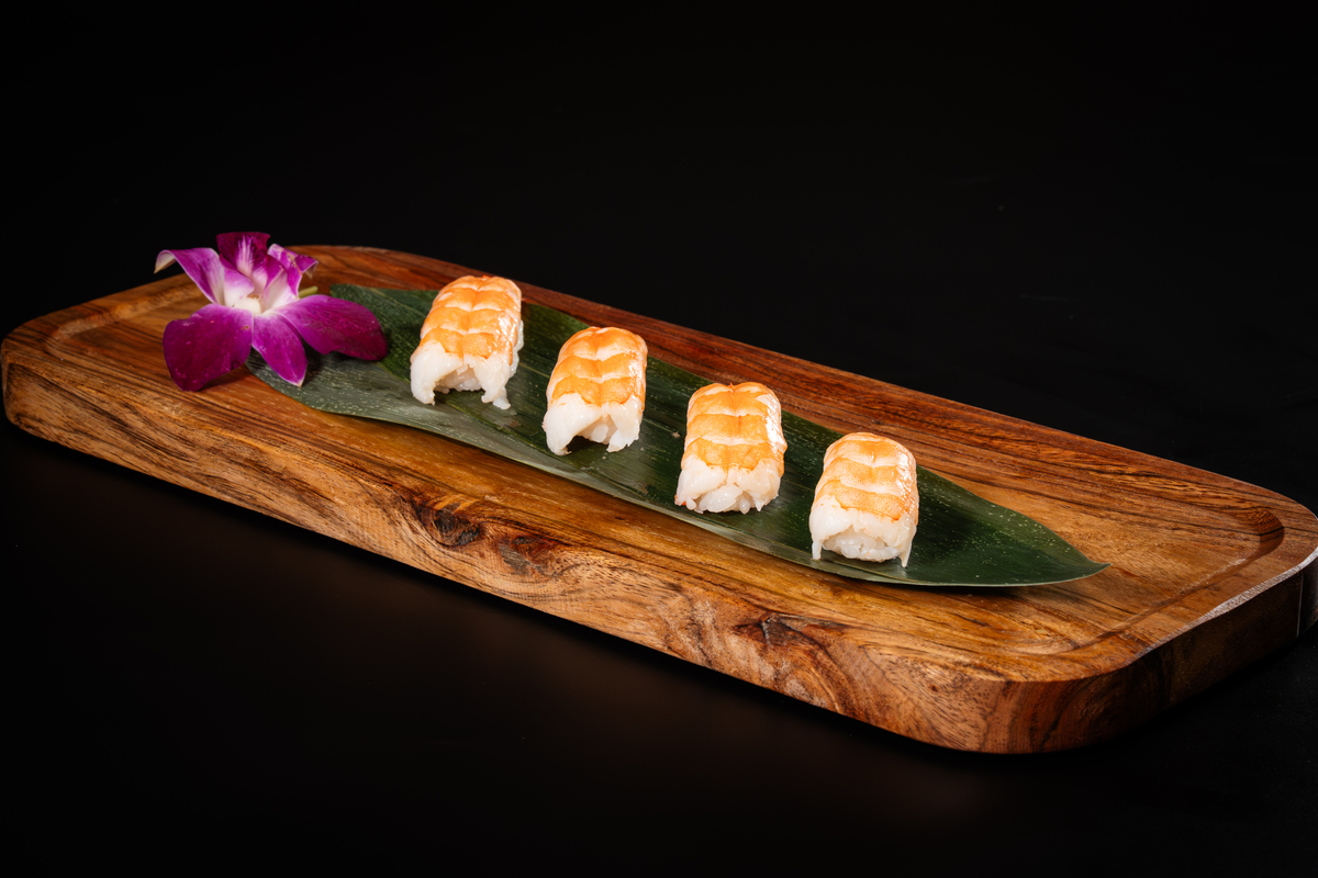 Four pieces of shrimp nigiri sushi are arranged on a green leaf atop a wooden tray, with a purple orchid garnish, set against a black background—just one tempting highlight from the Kingdom Sushi menu.