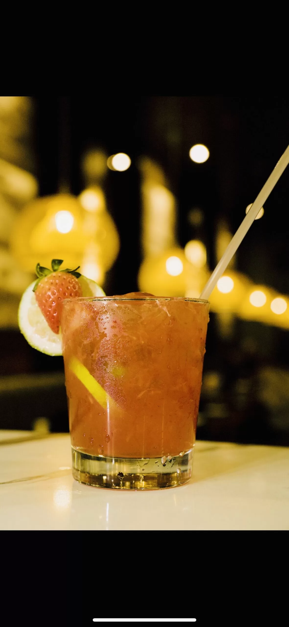 A cold, red Strawberry Lime Caipisake garnished with a slice of lime and a strawberry on the rim. The drink is served in a glass with a straw, resting on a white surface as warm, blurry lights create a cozy background ambiance.