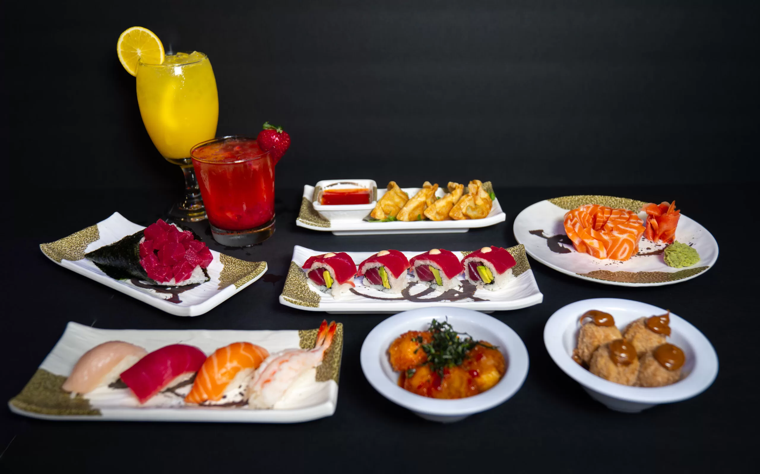 A variety of sushi, sashimi, appetizers, and two colorful drinks are arranged on white plates against a black background. The dishes include rolls, nigiri, fried items, and sliced raw fish.